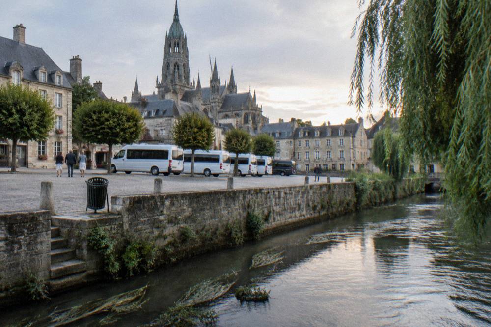 A stone square with parked minibuses beside the river in Bayeux
