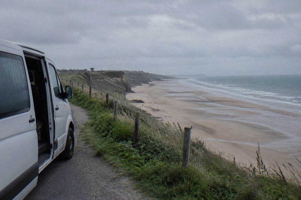 A guided tour van parked on a coastal road above a wide Normandy beach