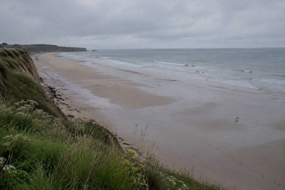 Wide empty sandy beach below grassy bluffs under a gray sky