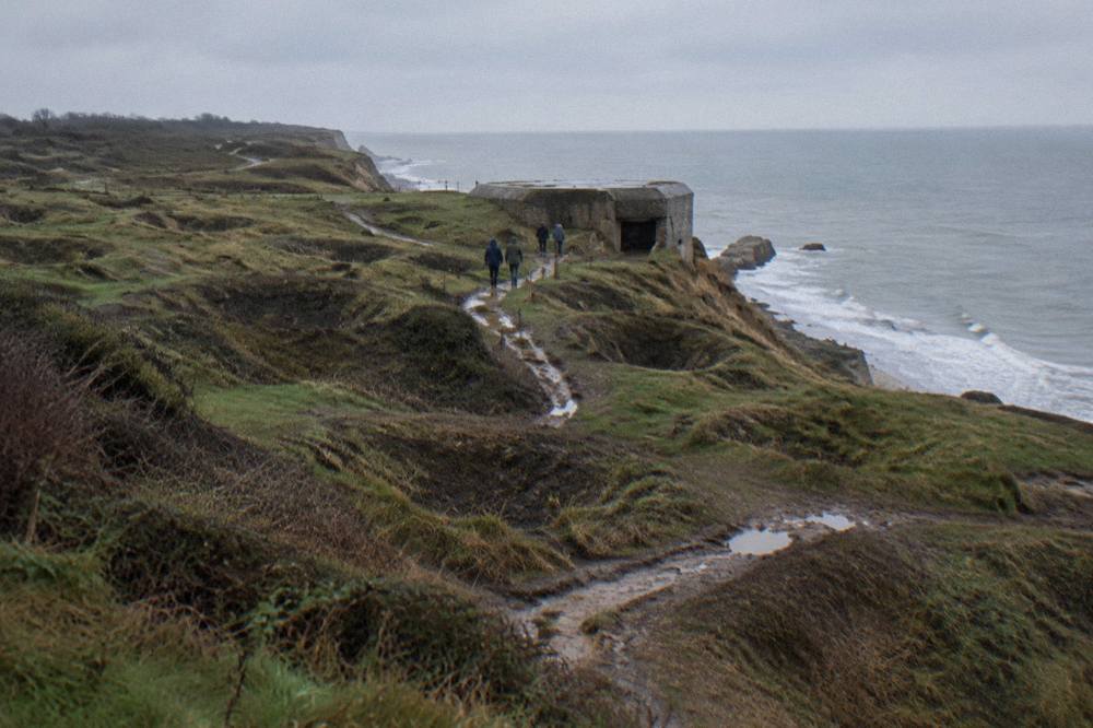 A worn footpath crossing grass toward a stone bunker on a cliff top above the sea