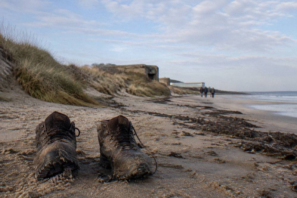 Walking shoes standing on damp sand beside a stretch of quiet beach below a grassy bluff
