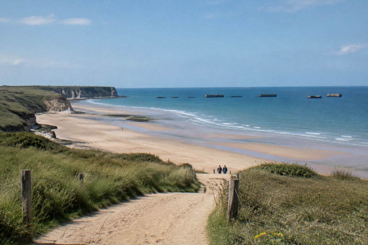 Wide sandy beach below low bluffs with remnants of a wartime harbor in the distance