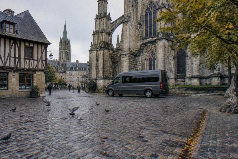 A minibus parked on a town square beside medieval stone buildings in Bayeux