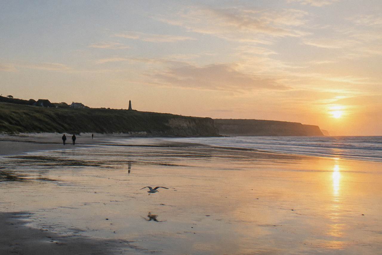 Wide view of Omaha Beach at low tide with the bluff and memorial markers along the sand