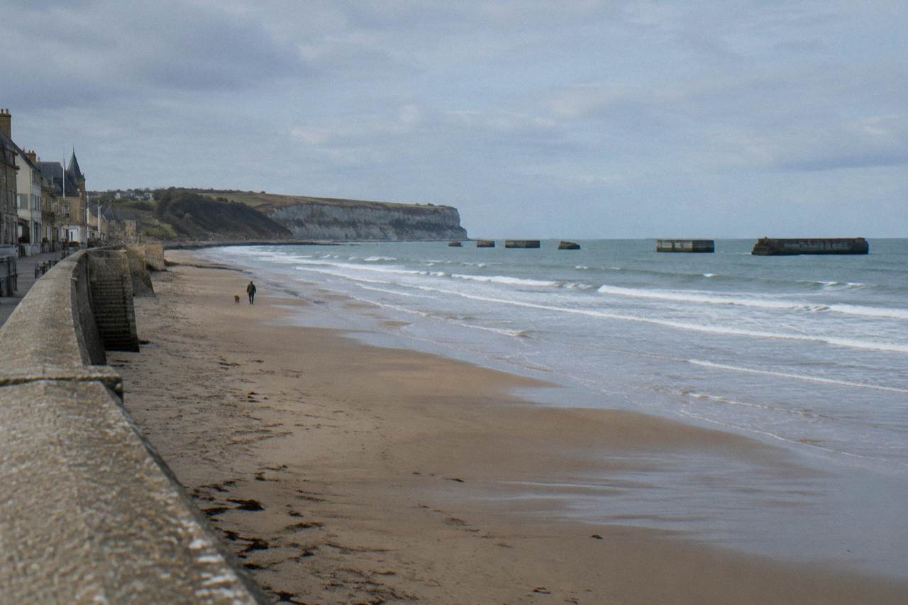 Wide sandy beach below grassy bluffs with remnants of a wartime harbour in the distance