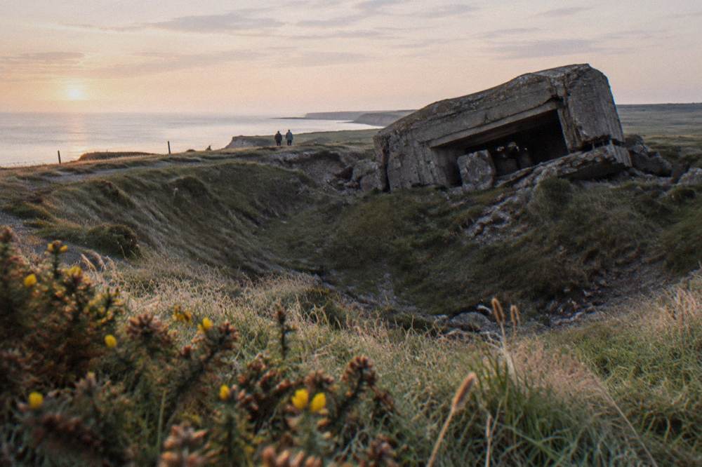 Grassy clifftop field pocked with large craters beside a concrete bunker ruin