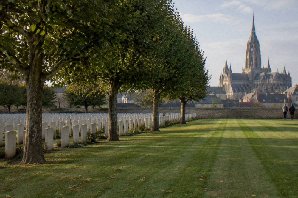Rows of headstones in a Commonwealth war cemetery with flat lawn and trees on a still afternoon