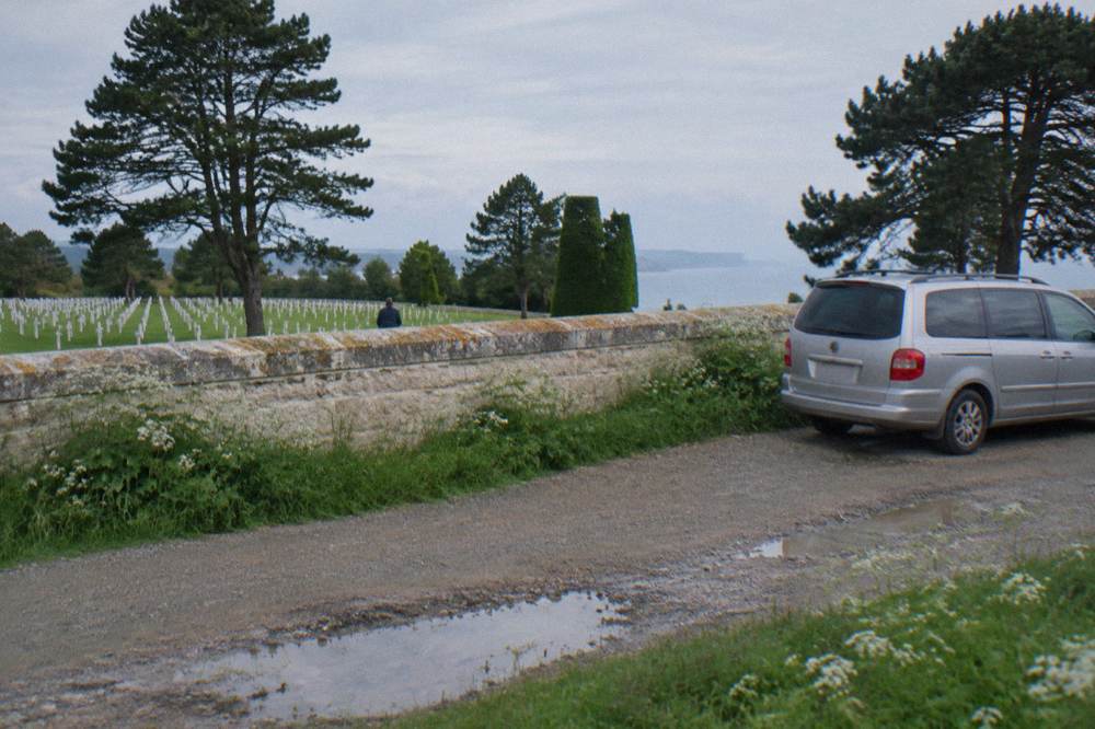 A minivan parked on a gravel lane near a stone memorial wall