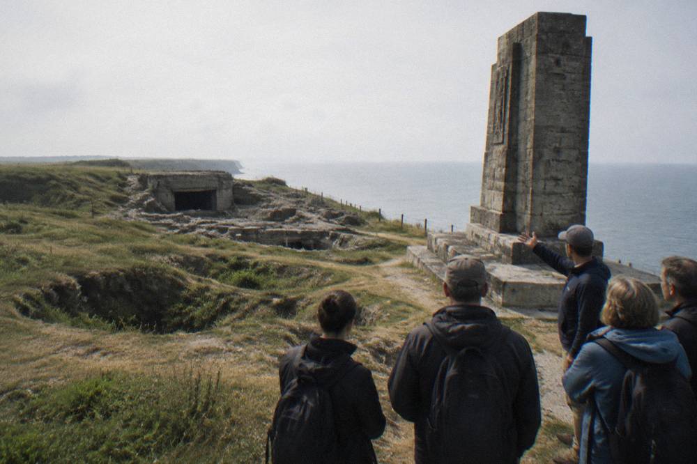Guide addressing a small group beside a stone memorial on a Normandy clifftop
