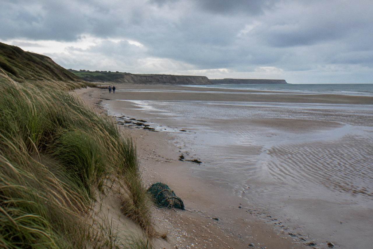 Wide sandy stretch of Omaha Beach at low tide with dune grass and calm gray sea