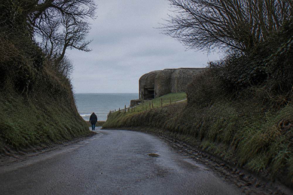 A quiet country road running between hedgerows toward the Normandy coast