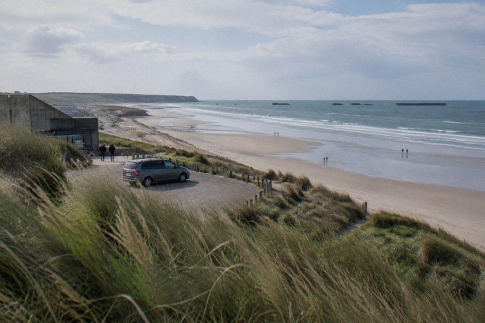 A shared tour minibus parked above a wide sandy Normandy landing beach