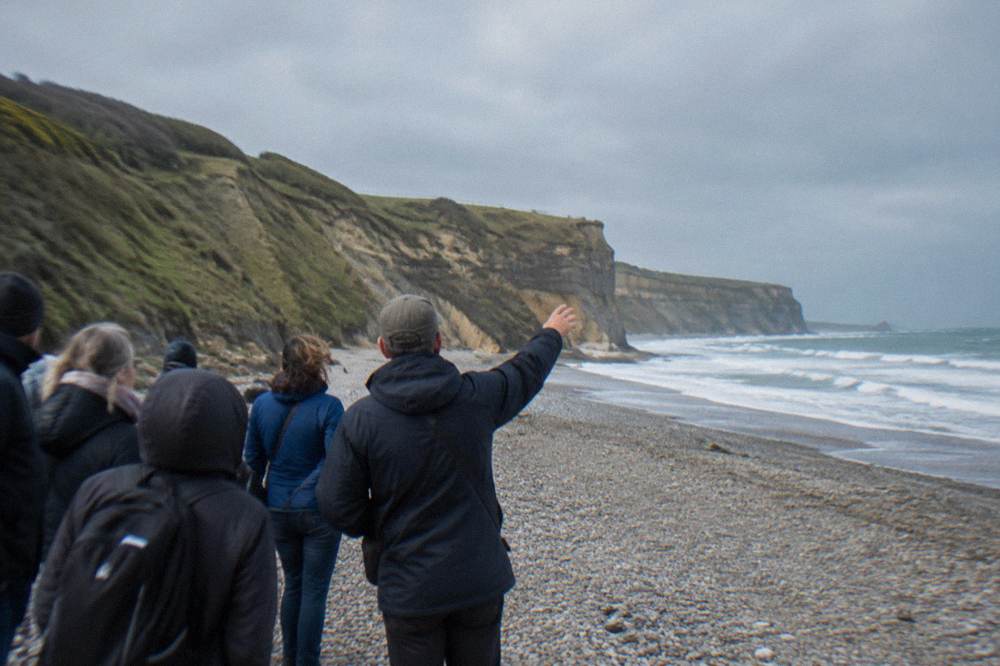 A guide speaking to a small cluster of visitors beside a stone memorial near Omaha Beach
