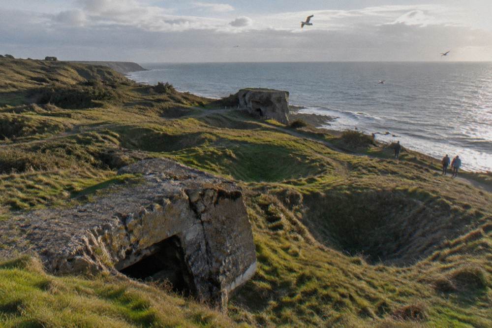 Wind-swept clifftop field pocked with large grass-grown craters and broken concrete bunkers