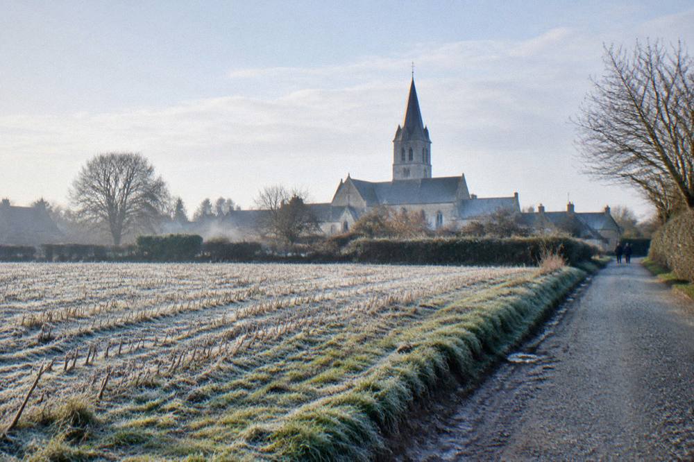 Country road through flat Norman farmland toward a stone church spire on the horizon