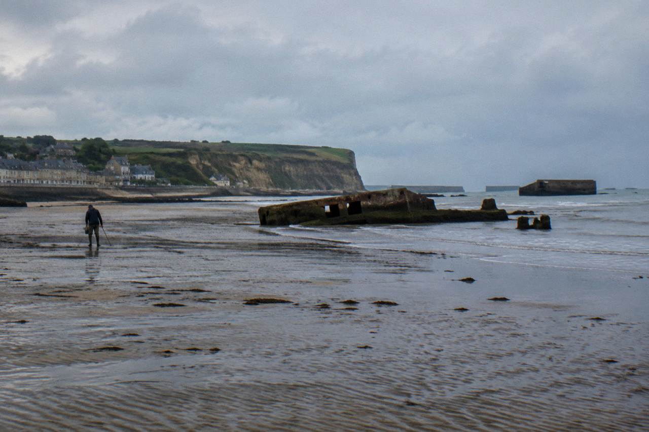 Wide sandy beach below low bluffs with remnants of wartime harbor structures along the shoreline