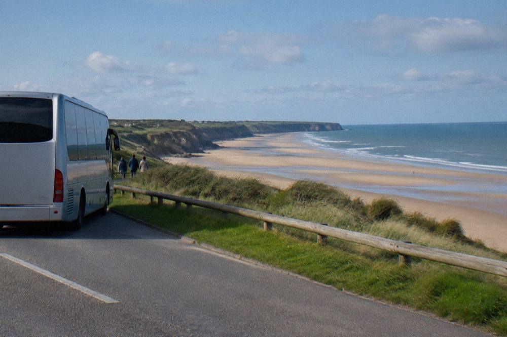 A touring coach parked on a coastal road above a wide Normandy beach