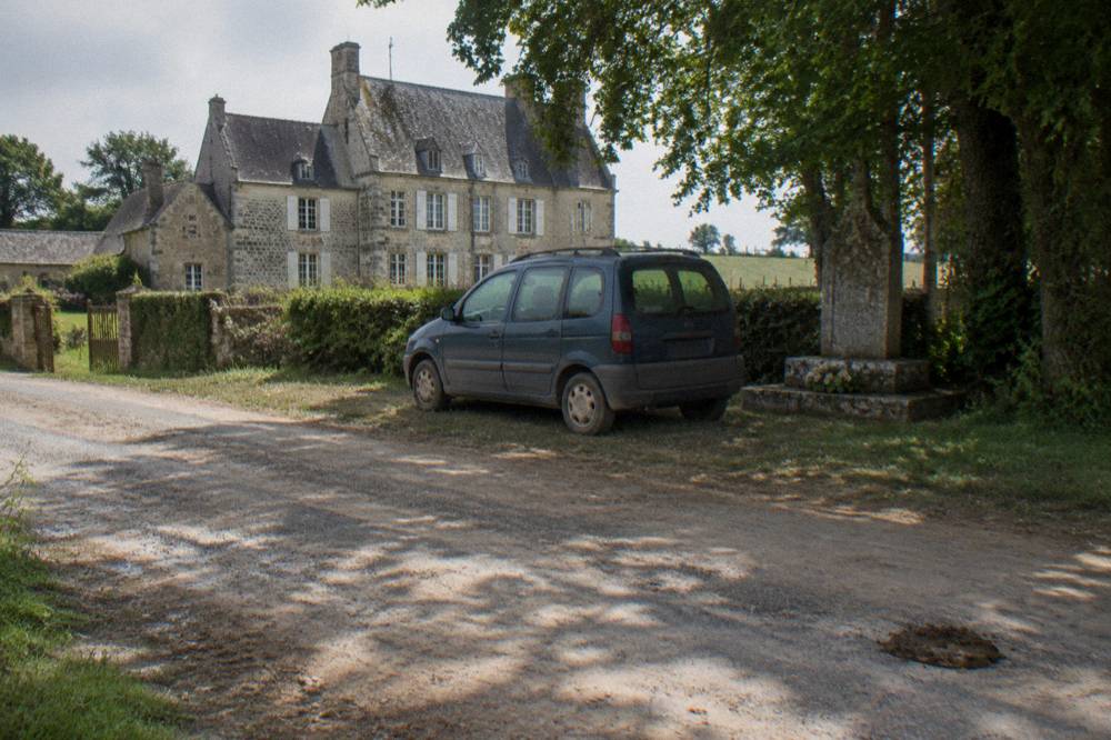 A small minivan parked beside a stone memorial on a quiet Normandy lane