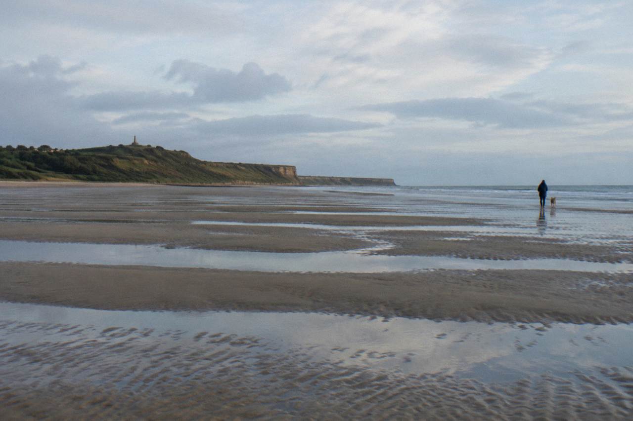 Wide view of Omaha Beach sand at low tide with the bluff and memorial visible in the distance